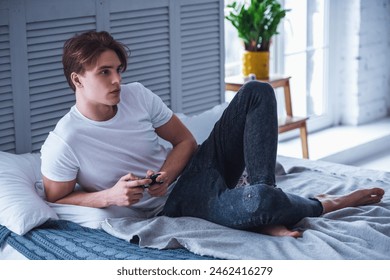 Handsome young man in casual clothes is playing video game console while sitting on his bed at home - Powered by Shutterstock