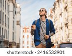 Handsome young man in casual clothing enjoying summer and wandering empty streets while walking outdoors with cup of coffee in hands. Solo trip city exploration