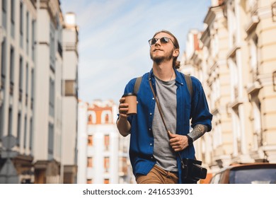 Handsome young man in casual clothing enjoying summer and wandering empty streets while walking outdoors with cup of coffee in hands. Solo trip city exploration - Powered by Shutterstock