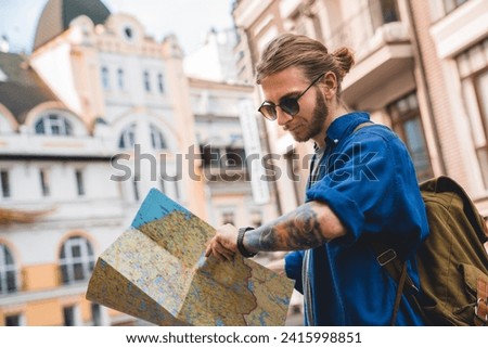 Handsome young man in casual clothing holding map and checking the time while standing on the city streets outdoors. Tourist finding the direction, way, route in a new city
