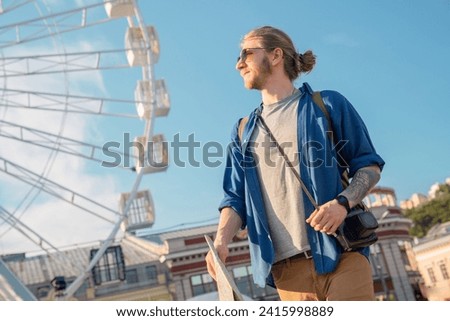 Handsome young man in casual clothing holding map and looking away while standing outdoors. Caucasian hipster walking around the new city checking directions route