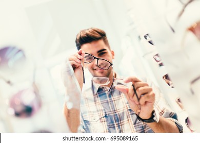 Handsome Young Man Choosing Eyeglasses Frame In Optical Store.