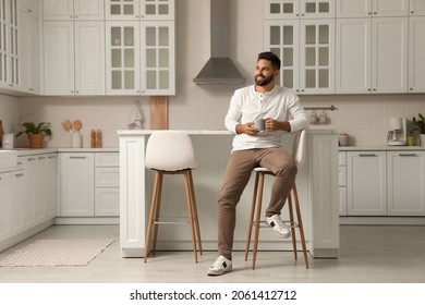 Handsome young man with cup of tea sitting on stool in kitchen - Powered by Shutterstock