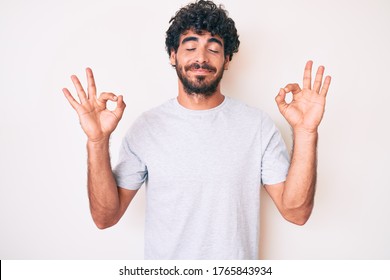 Handsome young man with curly hair and bear wearing casual tshirt relax and smiling with eyes closed doing meditation gesture with fingers. yoga concept. 