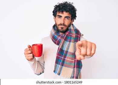 Handsome Young Man With Curly Hair And Bear Wearing Winter Scarf And Drinking A Cup Of Hot Coffee Pointing With Finger To The Camera And To You, Confident Gesture Looking Serious 