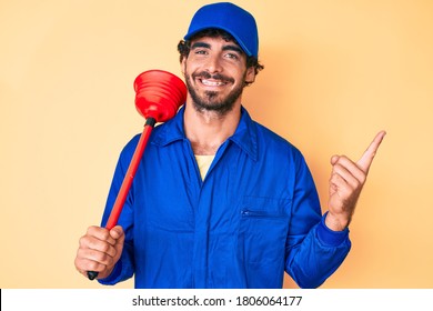 Handsome young man with curly hair and bear wearing plumber uniform holding toilet plunger smiling happy pointing with hand and finger to the side 