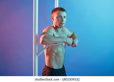 Handsome young man doing exercise on horizontal bar, guy posing topless, keeps fit, posing isolated over neon background, male looking aside with calm expression.