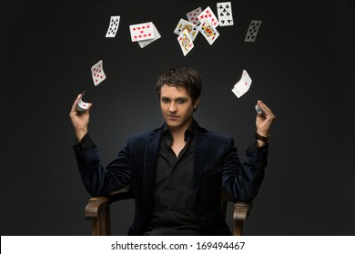 Handsome Young Man Dressed In Black Suit Juggling Playing Cards Over Black Background