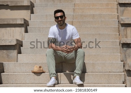 Handsome young man dressed in casual clothes and sunglasses sitting on the stairs of the park. The man has a hat and enjoys the sunny day in spring. Travel and holiday concept