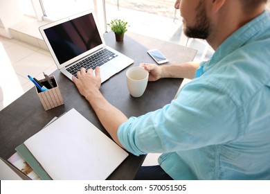 Schöner junger Mann, der Kaffee trinkt, während er mit Laptop zu Hause arbeitet