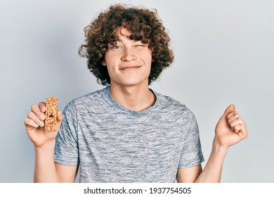 Handsome Young Man Eating Protein Bar As Healthy Energy Snack Screaming Proud, Celebrating Victory And Success Very Excited With Raised Arm 