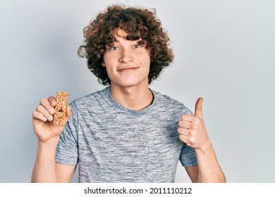 Handsome Young Man Eating Protein Bar As Healthy Energy Snack Smiling Happy And Positive, Thumb Up Doing Excellent And Approval Sign 