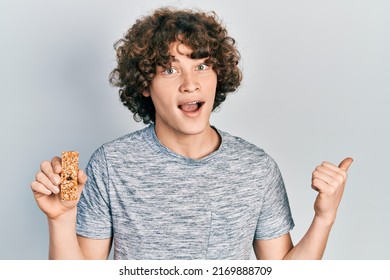 Handsome Young Man Eating Protein Bar As Healthy Energy Snack Pointing Thumb Up To The Side Smiling Happy With Open Mouth 