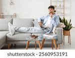 Handsome young man eating sweet chocolate bar with modern laptop and sitting on sofa in living room