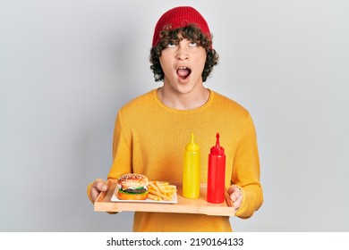 Handsome young man eating a tasty classic burger with ketchup and mustard angry and mad screaming frustrated and furious, shouting with anger looking up.  - Shutterstock ID 2190164133