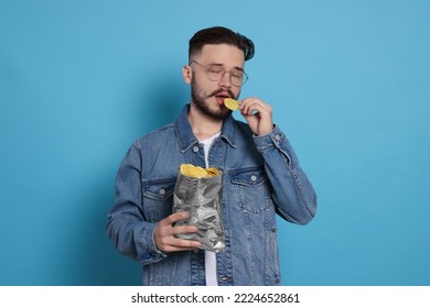 Handsome young man eating tasty potato chips on light blue background