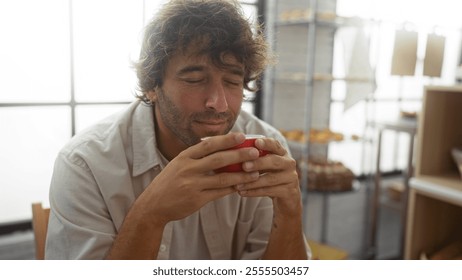 Handsome young man enjoying coffee indoors in a bakery, savoring the aroma while seated in a cozy, sunlit space. - Powered by Shutterstock