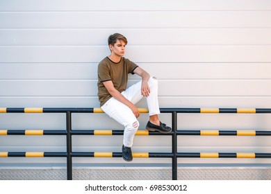 handsome young man in a fashionable classic T-shirt and white pants with shoes sits near a metal glossy wall