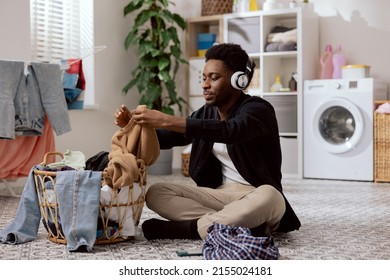 Handsome Young Man Folds Clean Clothes Sorts Laundry Before Putting It In Washing Machine College Student Sits On Bathroom Floor Listening To Music On Wireless Headphones.