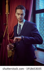 Handsome Young Man In A Formal Suit Is Looking At A Watch And Holding A Glass Of Whiskey In A Luxury Classic Interior. Business Style. Men's Club.