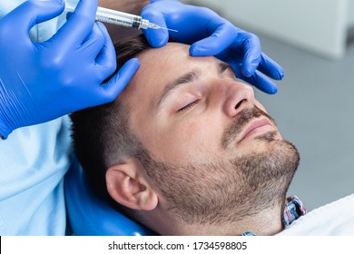 Handsome young man is getting a rejuvenating facial injections. He is sitting calmly at clinic. The expert beautician is filling male wrinkles by hyaluronic acid.