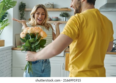 Handsome young man giving a bunch of flowers to his girlfriend while standing at the kitchen - Powered by Shutterstock