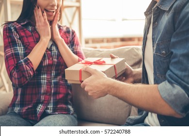 Handsome young man is giving a present to his lovely girlfriend while they are sitting on couch at home - Shutterstock ID 572240788