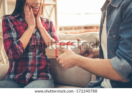 Handsome young man is giving a present to his lovely girlfriend while they are sitting on couch at home