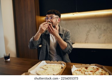 A handsome young man in glasses is enjoying pizza in his kitchen. Unbelievably enjoys the taste. - Shutterstock ID 2256055373