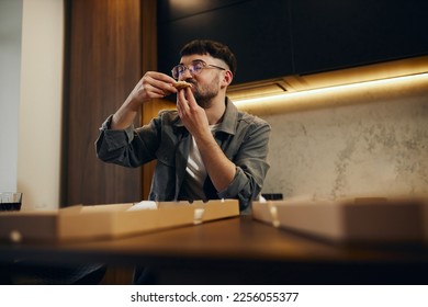 A handsome young man in glasses is enjoying pizza in his kitchen. Unbelievably enjoys the taste. - Shutterstock ID 2256055377