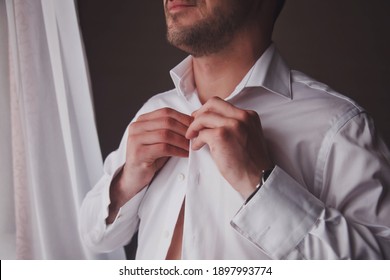 Handsome Young Man Groom Dresses White Shirt At Window In Hotel Room, Smile With Sure Look Himself. Cute Guy Is Getting Ready To Meet Bride. Concept Of Happiness And Luxury Married. Copy Space