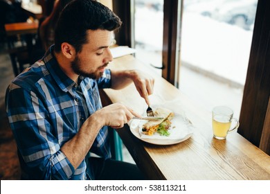 Handsome young man having lunch in cosy restaurant or cafe. He is sitting alone near in front of window. Concentrated guy is using knife and fork to cut his sandwich - Powered by Shutterstock