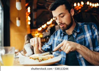 Handsome young man having lunch in elegant restaurant alone. Attractive guy is cutting his tasty sandwich with fork and knife. He is delighted because soon he will try this delicious dish - Powered by Shutterstock