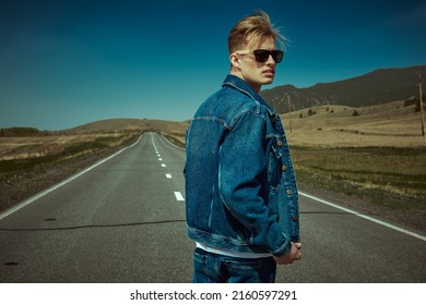 Handsome Young Man Hitchhiker In Denim Clothes And Black Sunglasses Standing On A Road Over Picturesque Landscape. Denim Fashion. Road Adventures, Travelling.