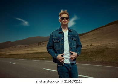 Handsome Young Man Hitchhiker In Denim Clothes And Black Sunglasses Standing On A Road Over Picturesque Landscape. Denim Fashion. Road Adventures, Travelling.