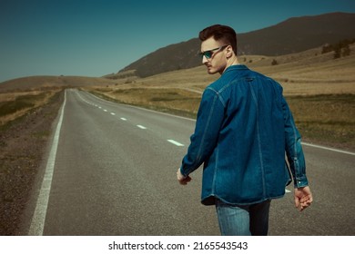 Handsome Young Man Hitchhiker In Denim Clothes And Black Sunglasses Walking On A Road Over Picturesque Landscape. Denim Fashion. Road Adventures, Travelling.