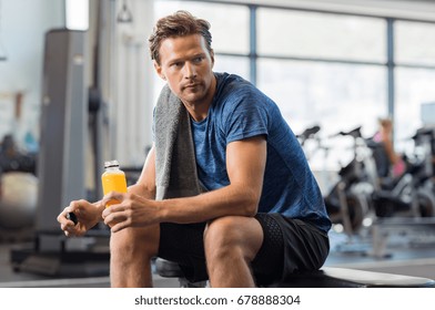 Handsome young man holding bottle of fresh orange juice while resting at gym.Thoughtful fit man sitting alone holding a bottle of energy drink. Guy take break after fitness exercise on bench.