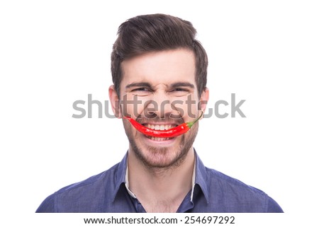 Handsome young man is holding chili pepper in his teeth while standing against white background.
