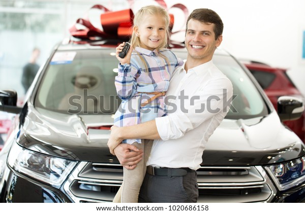 Handsome young man holding his adorable daughter\
while she is showing car keys to their newly bought auto posing at\
the local dealership consumerism buying purchase rental leasing\
service family