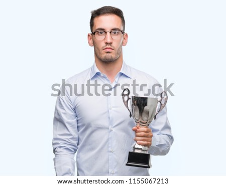 Handsome young man holding trophy with a confident expression on smart face thinking serious