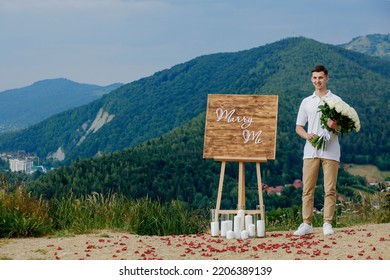 Handsome young man holding white roses, smiling against mountain landscape background. Marry me