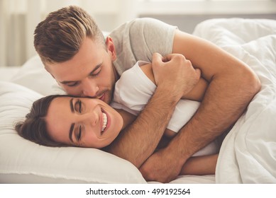 Handsome young man is hugging and kissing his beautiful smiling wife in cheek while they both are lying in bed in the morning - Powered by Shutterstock