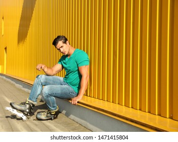 Handsome young man with inline roller skates sitting near yellow building, space for text