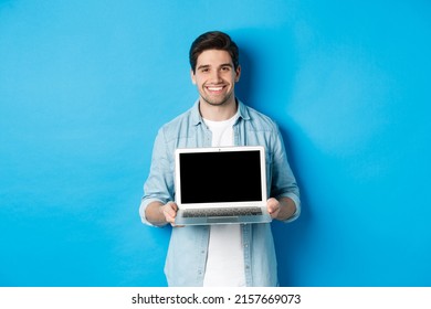 Handsome Young Man Introduce Product On Laptop Screen, Showing Computer And Smiling, Standing Over Blue Background