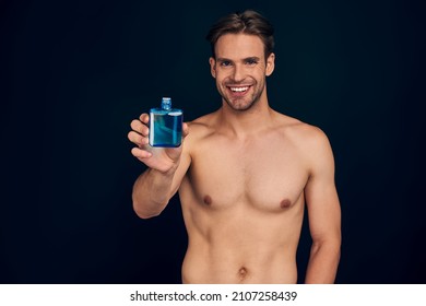Handsome young man isolated. Portrait of shirtless muscular man is standing on dark blue background and using face aftershave lotion. Men care concept