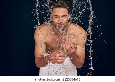 Handsome young man isolated. Portrait of shirtless muscular man is standing on grey background and washing oneself. Spraying water into face. Men care concept
