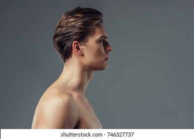 Handsome Young Man Isolated. Portrait Of Shirtless Muscular Man Is Standing On Grey Background Turned In Profile.