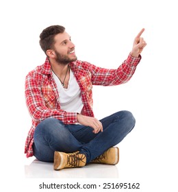 Handsome Young Man In Jeans And Lumberjack Shirt Sitting On Floor With Legs Crossed And Pointing Up. Full Length Studio Shot Isolated On White.