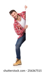 Handsome young man in jeans and lumberjack shirt standing behind white banner and smiling. Full length studio shot isolated on white.