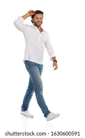 Handsome Young Man In Jeans, Sneakers And White Shirt Is Walking, Holding Sunglasses On His Head, Looking At Camera And Smiling. Side View. Full Length Studio Shot Isolated On White.